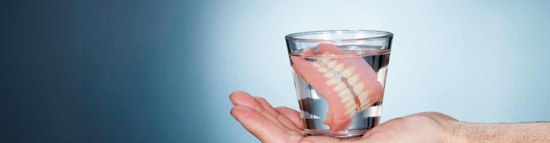 Hand holding a clear glass of water with a full set of removable dentures soaking inside, illustrating proper denture cleaning and overnight storage.